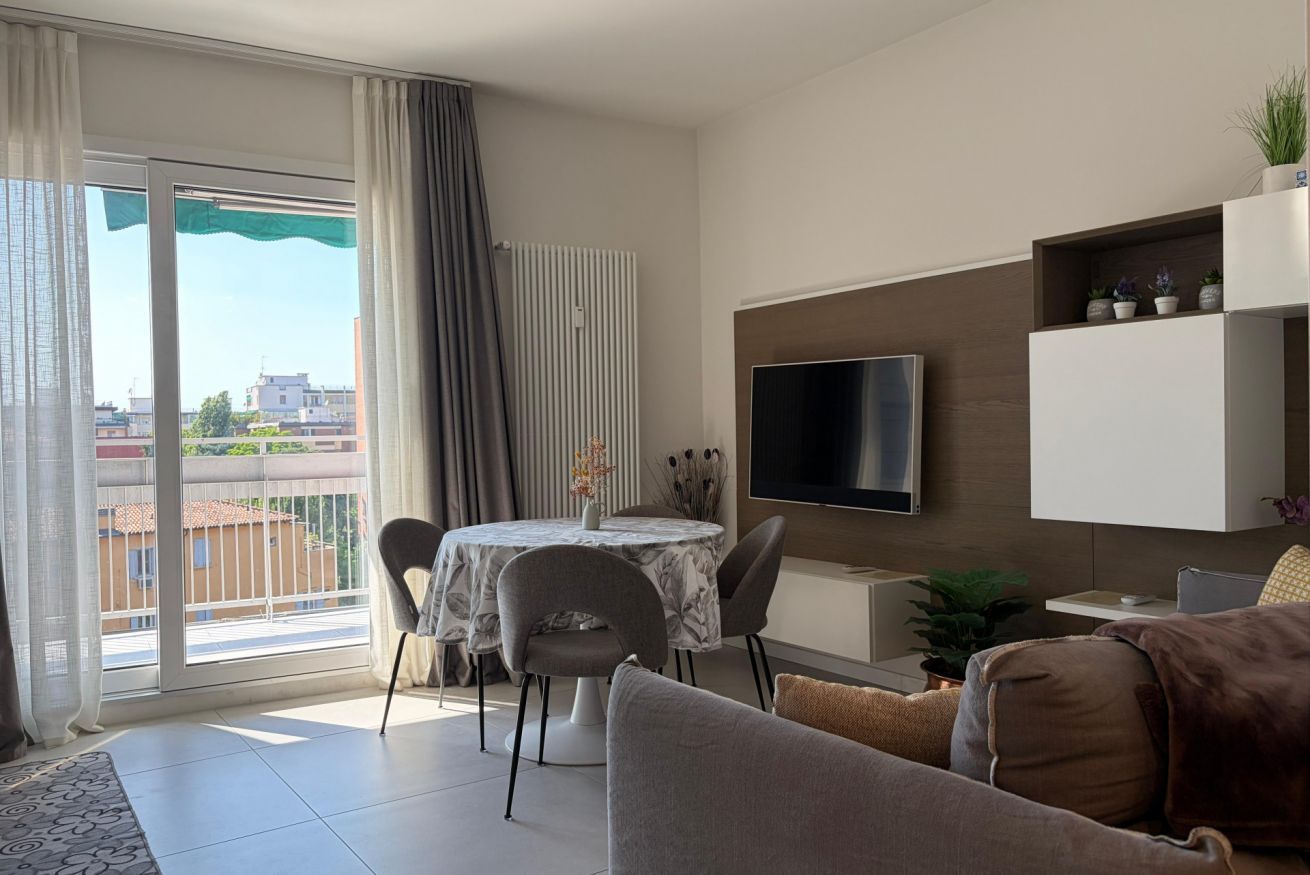 Un salon moderne doté d’un téléviseur à écran plat, d’un coin repas avec une table et des chaises, d’un canapé et d’un balcon agrémenté de rideaux.