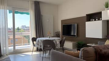 Un salon moderne doté d’un téléviseur à écran plat, d’un coin repas avec une table et des chaises, d’un canapé et d’un balcon agrémenté de rideaux.