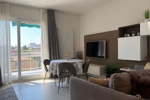 Un salon moderne doté d’un téléviseur à écran plat, d’un coin repas avec une table et des chaises, d’un canapé et d’un balcon agrémenté de rideaux.