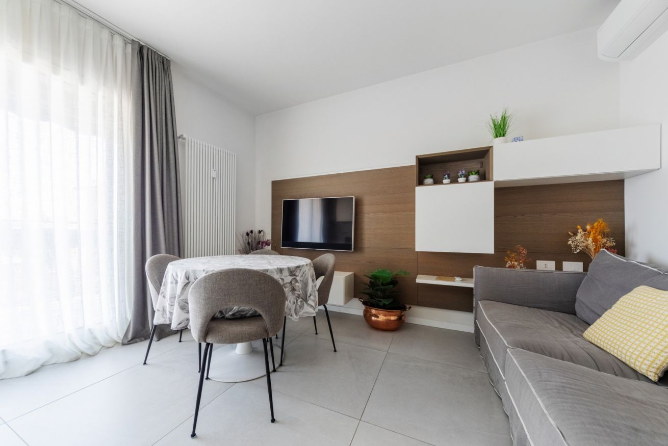 Een moderne woonkamer met een eethoek, een flatscreen televisie aan de muur, een grijze bank en een kleine tafel met stoelen.