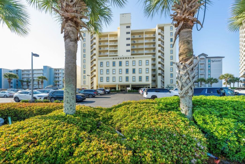 A multi-story hotel with a central entrance, surrounded by a parking lot and palm trees.