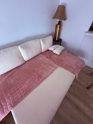 Ein Bett mit einer rosa Tagesdecke, einem weißen Kopfkissen und einem beigen Kissen. Auf einem kleinen Tisch neben dem Bett steht eine Lampe.