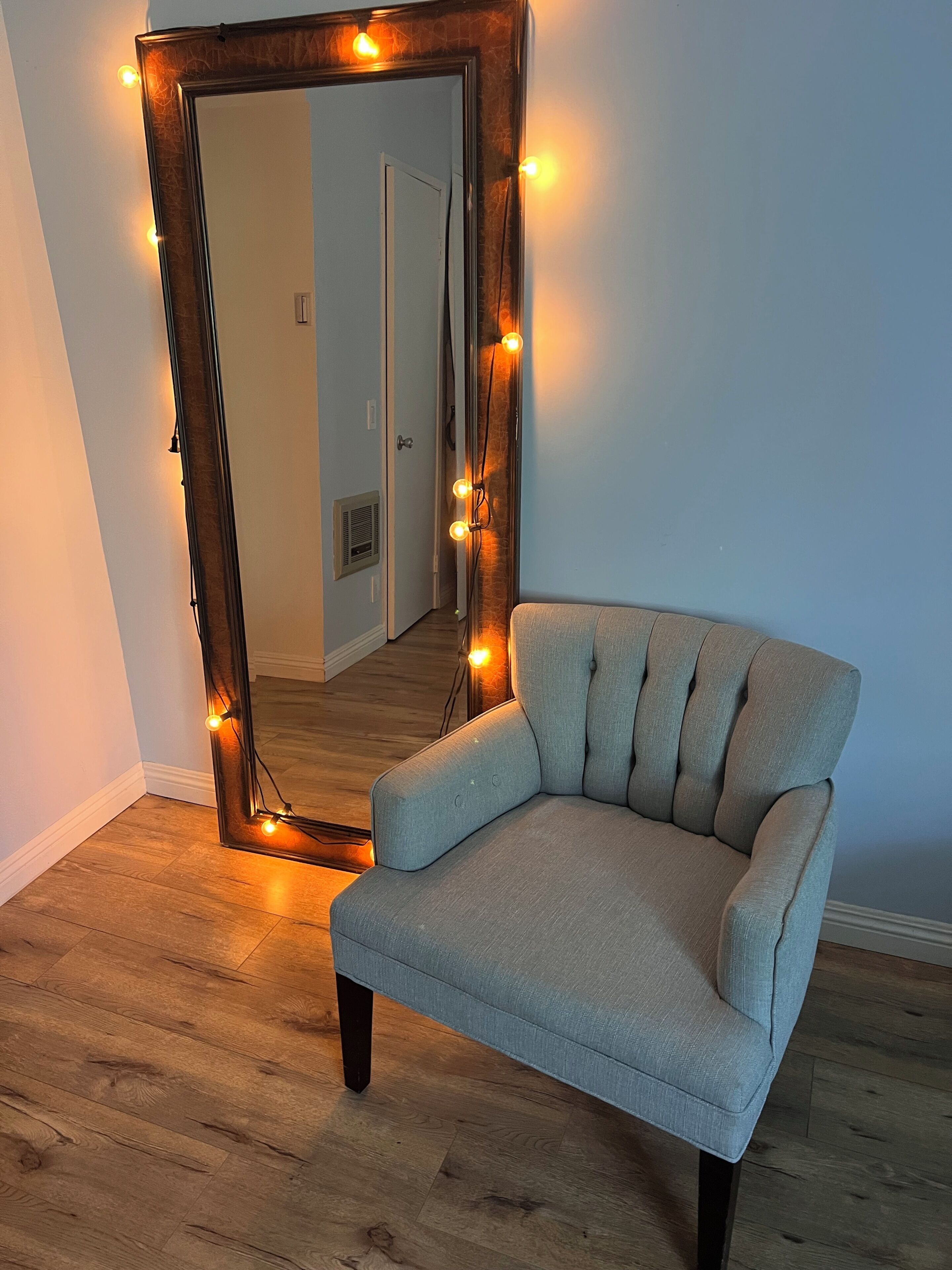 Una habitació amb un mirall gran, un divà a color clar, terres de fusta i una porta al fons.
