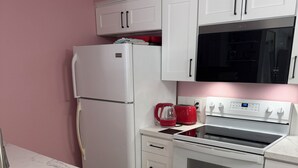 Una cocina con armarios blancos, un refrigerador de acero inoxidable, una tetera roja, un microondas y un horno empotrado.