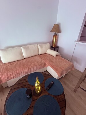 Ein modernes Wohnzimmer mit einem Ecksofa, einer Lampe, einem kleinen Tisch mit Dekoration und einer Fernbedienung.