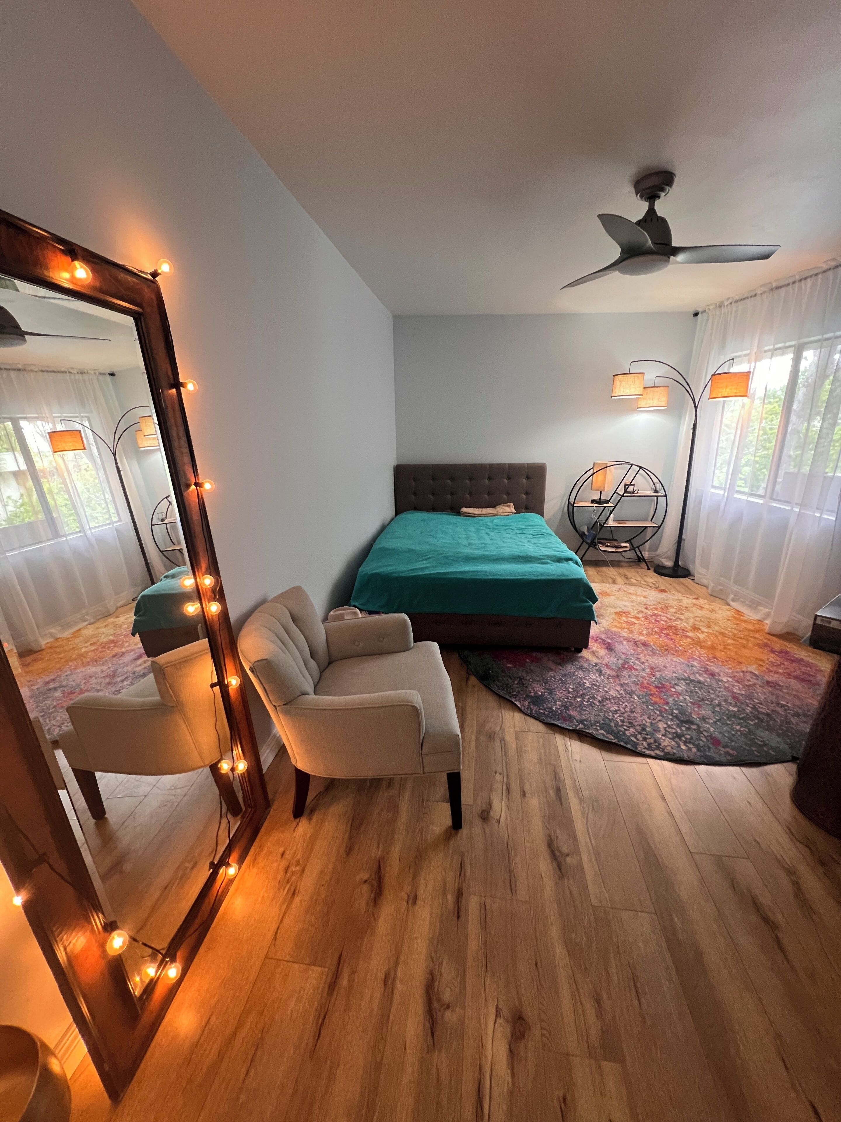 A bedroom with a bed, a chair, a mirror, a ceiling fan, and a window.
