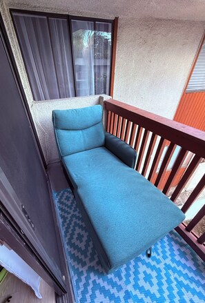 Ein kleines, zusammenklappbares Sofa mit blauer Sitzfläche, platziert auf einem Balkon mit Holzgeländer und einem gemusterten Teppich.