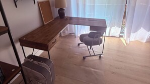 Una habitación con un escritorio de madera, una silla, un ventilador de pie, un estante con un marco de fotos y una ventana con cortinas translúcidas.