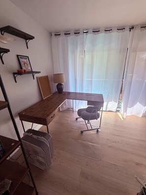Ein Zimmer mit einem Holztisch, einem Stuhl, einem Standventilator, einem Regal mit einem Bilderrahmen und einem Fenster mit durchsichtigen Vorhängen.