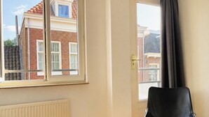 Ein Zimmer mit Heizkörper, einem schwarzen Stuhl und einem Fenster mit Blick auf ein Backsteingebäude.