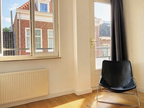 Ein Zimmer mit Heizkörper, einem schwarzen Stuhl und einem Fenster mit Blick auf ein Backsteingebäude.
