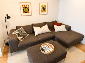 Ein braunes Ecksofa mit drei Kissen, ein kleiner Holztisch mit Tablett und zwei gerahmte Kunstwerke an der Wand.