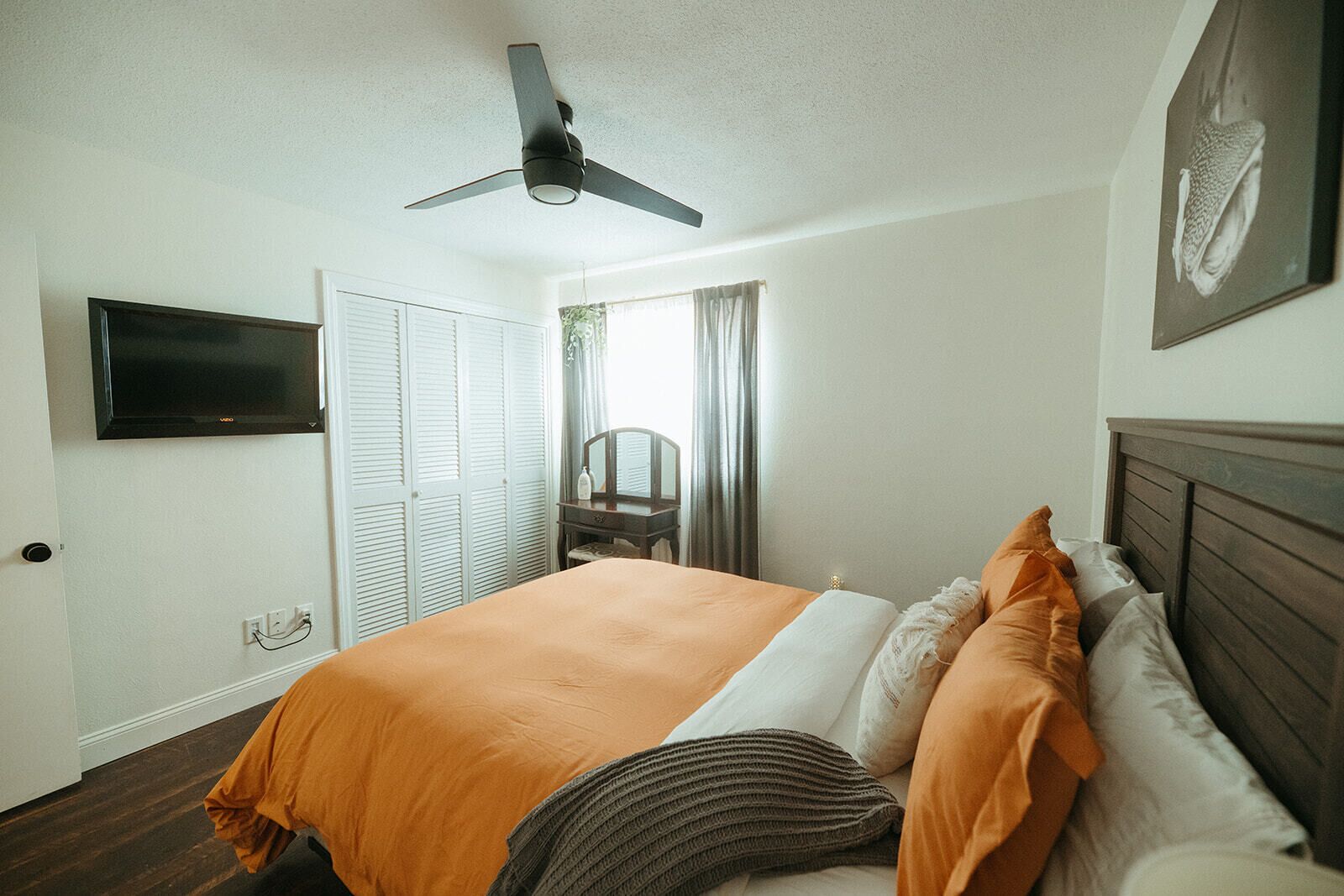 Ein Schlafzimmer mit einem Bett, einem Deckenventilator, einem an der Wand befestigten Fernseher und einem Fenster mit Vorhängen.