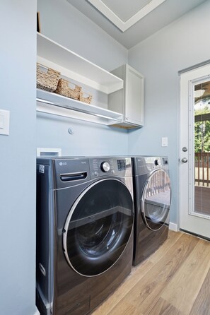 A laundry area with two front-loading LG washing machines, a white shelf above, and a door leading to an outdoor area. - Modern Retreat in Downtown Tulsa: Sleeps 7 (Tulsa)