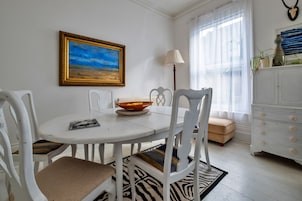 A dining area with a white table, chairs, a bowl of fruit, a framed painting, and a lamp.