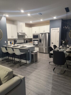 A modern kitchen with white cabinets, stainless steel appliances, and a dining area with a glass table and black chairs. - Perfect 4-bedroom home in Reunion Village West Kissimmee (Kissimmee)