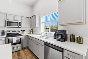 A modern kitchen with stainless steel appliances, white cabinetry, and a large window with blinds. - Curated Comfort in the Treasure Valley (Boise)