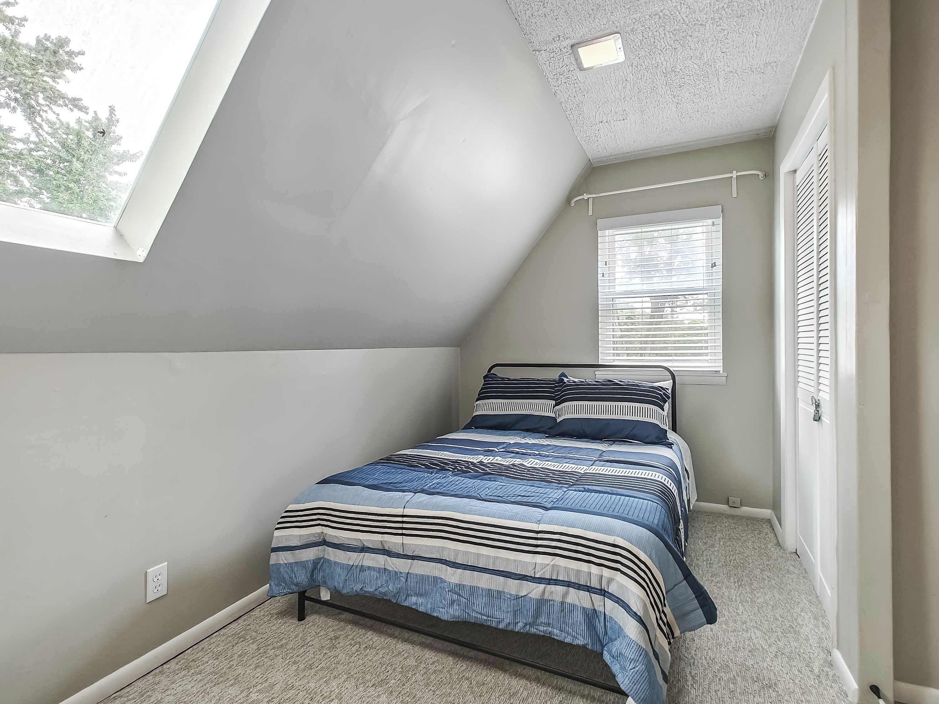 A bedroom with a sloped ceiling, a window with blinds, a bed with striped bedding, and a carpeted floor.