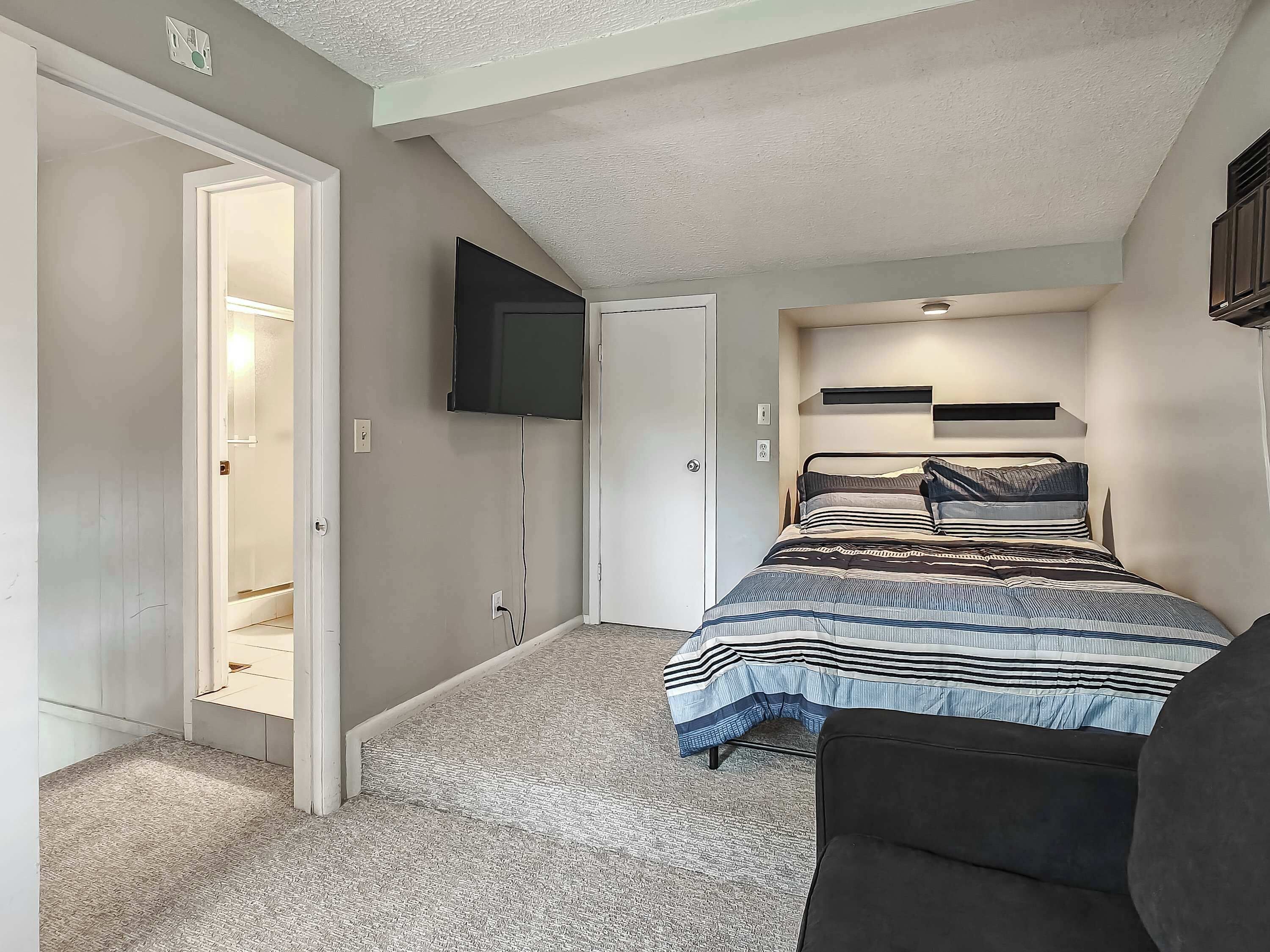 Ein Schlafzimmer mit einem Bett, einem an der Wand befestigten Fernseher, einer zur Garderobe führenden Tür und einem Teppichboden.