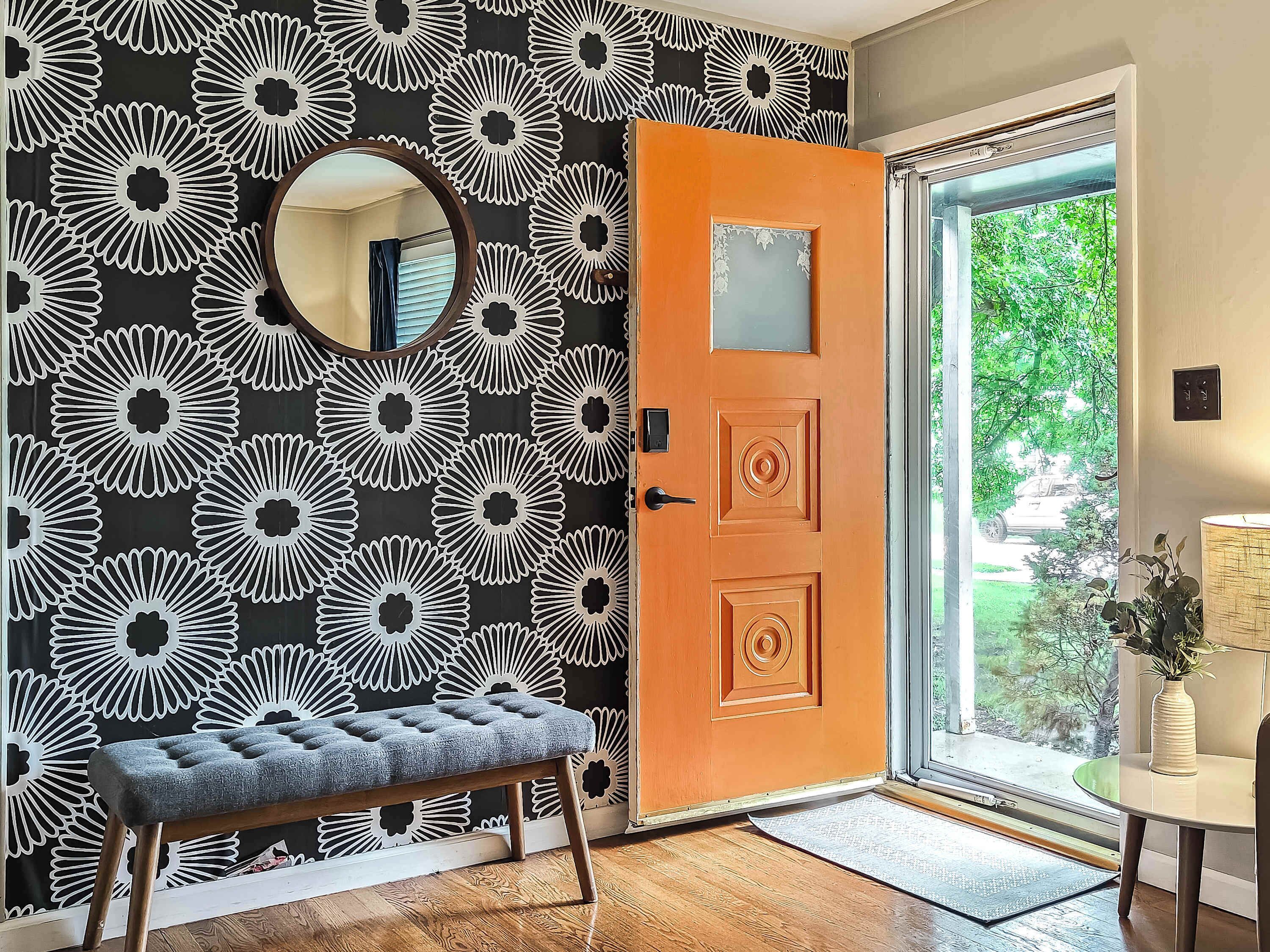 Quarto com papel de parede floral em preto e branco, um banco de madeira, um espelho redondo e uma porta com painel de vidro.