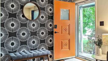 Quarto com papel de parede floral em preto e branco, um banco de madeira, um espelho redondo e uma porta com painel de vidro.
