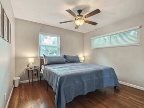 Un dormitorio con cama, ventilador de techo, dos mesitas de noche con lámparas y una ventana con persianas.