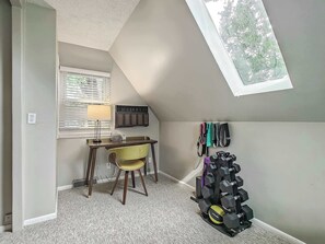Una habitación con techo inclinado, un escritorio con silla, una lámpara, un radiador y un soporte de pesas montado en la pared.