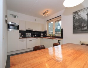 A modern kitchen with white cabinets, a wooden dining table, and a window with a view of a house. - 6-guest Deluxe Beach Villa With Sauna (Makkum)