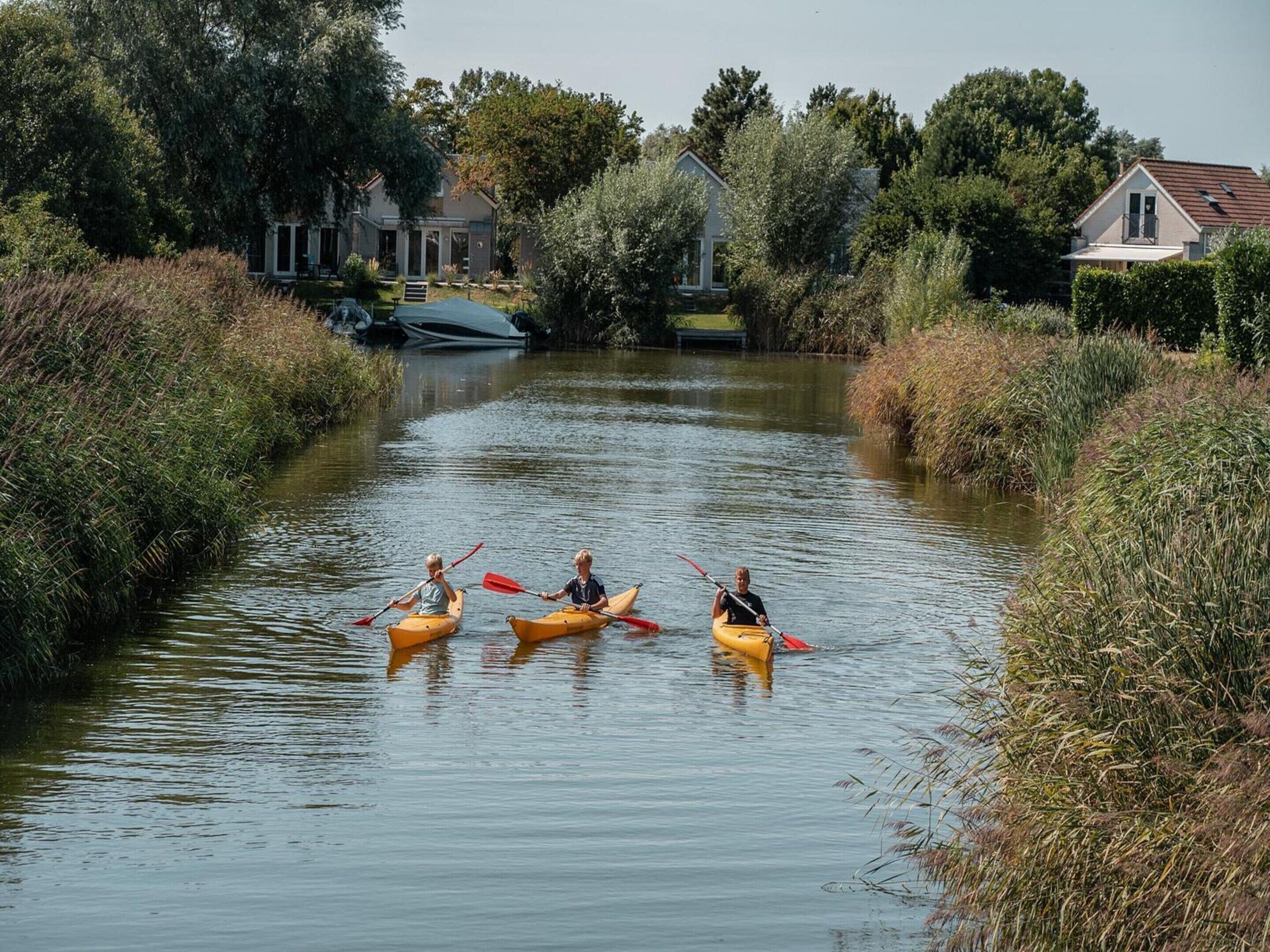 Drei Kajaks auf einem ruhigen Fluss, umgeben von Vegetation und Häusern.