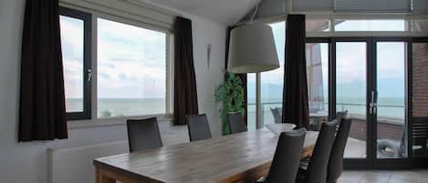 A dining area with a large table and chairs, large windows with curtains, a potted plant, and a view of the sea.