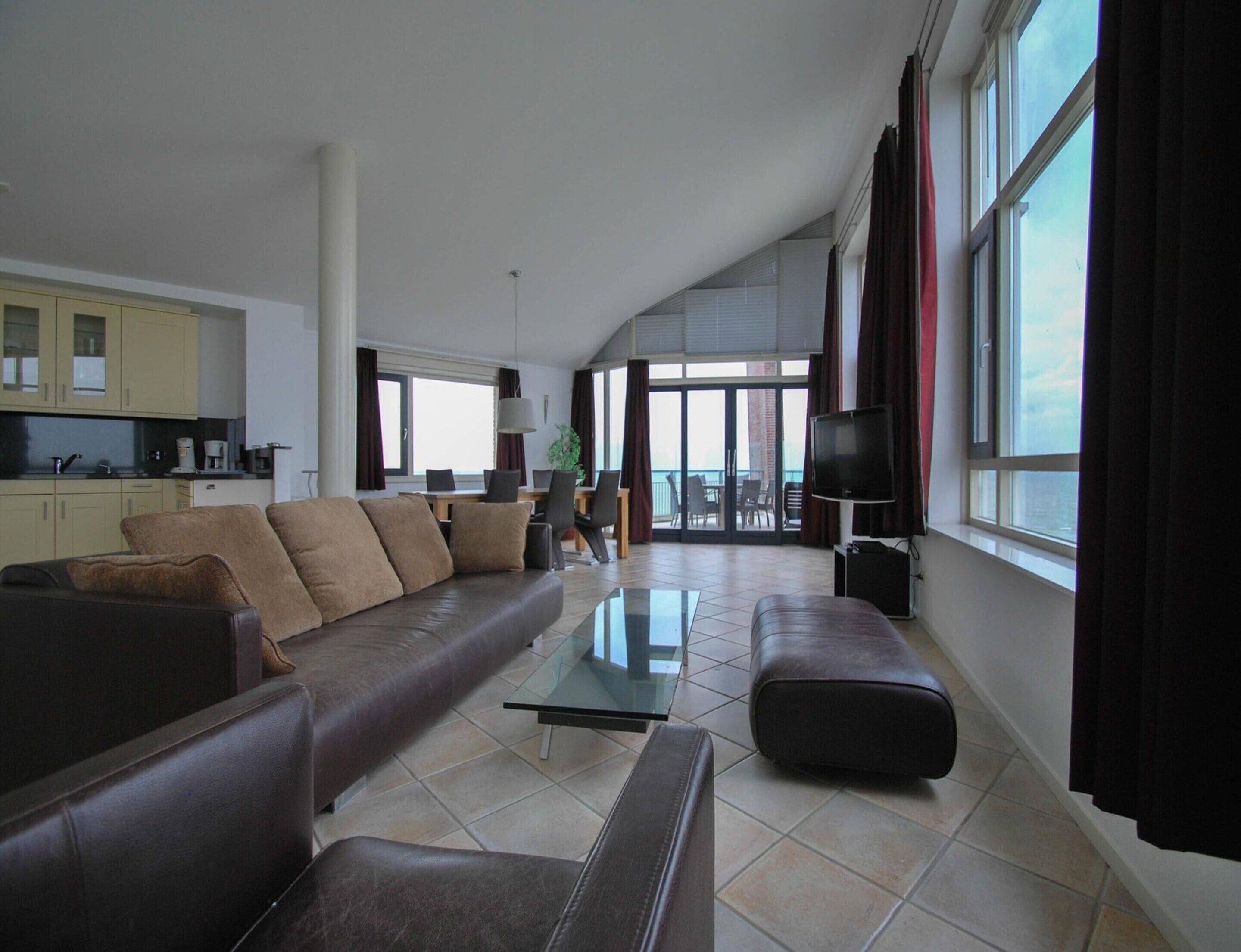 A spacious living area with a sofa, ottoman, and a glass coffee table. There is a kitchen area with cabinets and a dining table. Large windows provide natural light and a view outside.