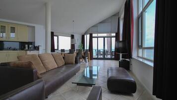 A spacious living area with a sofa, ottoman, and a glass coffee table. There is a kitchen area with cabinets and a dining table. Large windows provide natural light and a view outside.