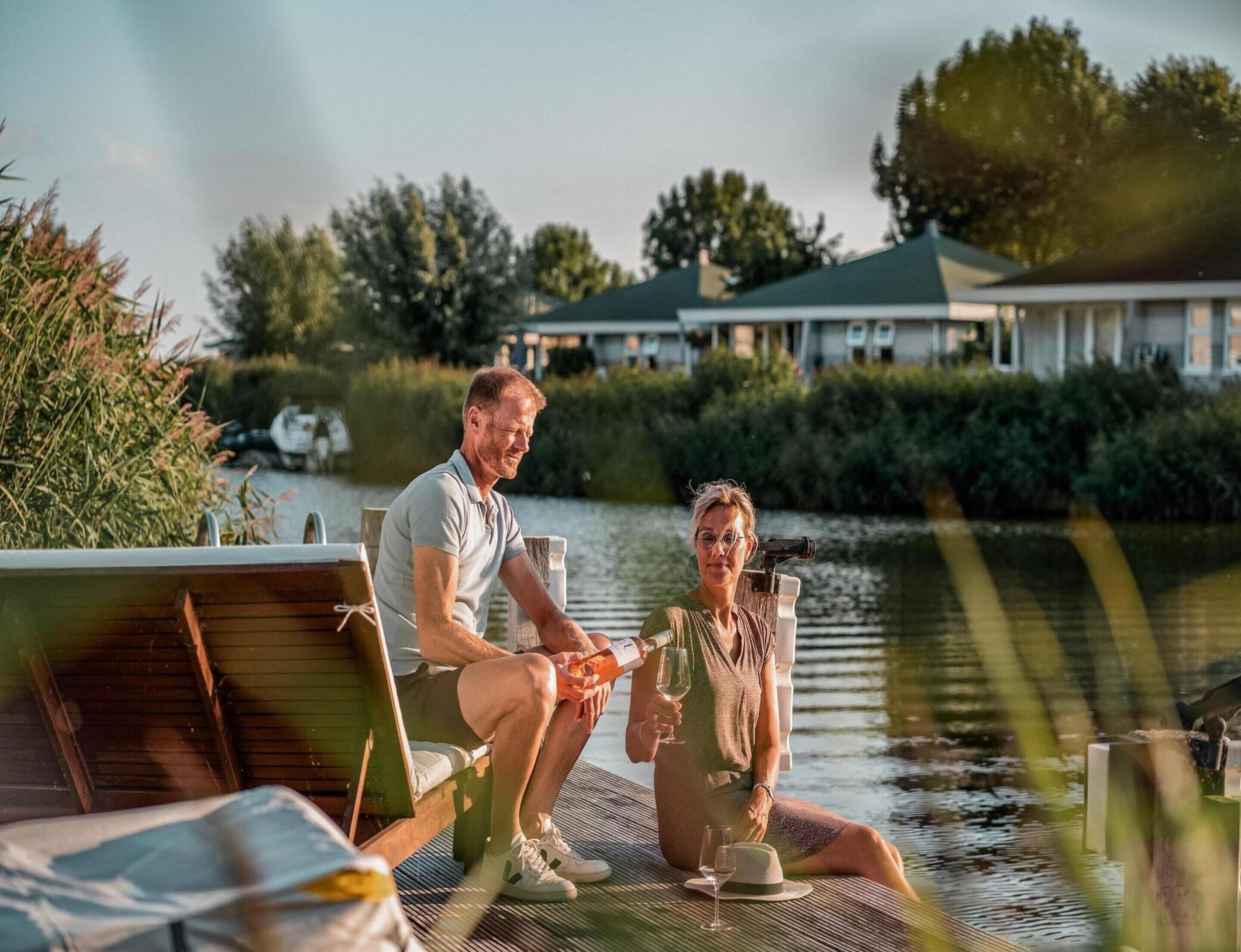 Two people seated on a dock with a body of water and greenery in the background.