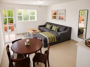 A living room with a grey sofa, a coffee table, and a dining table with chairs. There are two windows with blinds, a mirror, and framed pictures on the wall. - The Lighthouse Suite Umhlanga (uMhlanga)