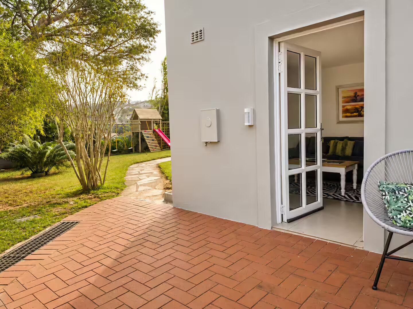 A patio area with a brick floor, a white door leading to a room, and a garden with a play structure.
