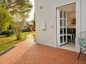 A patio area with a brick floor, a white door leading to a room, and a garden with a play structure. - The Lighthouse Suite Umhlanga (uMhlanga)