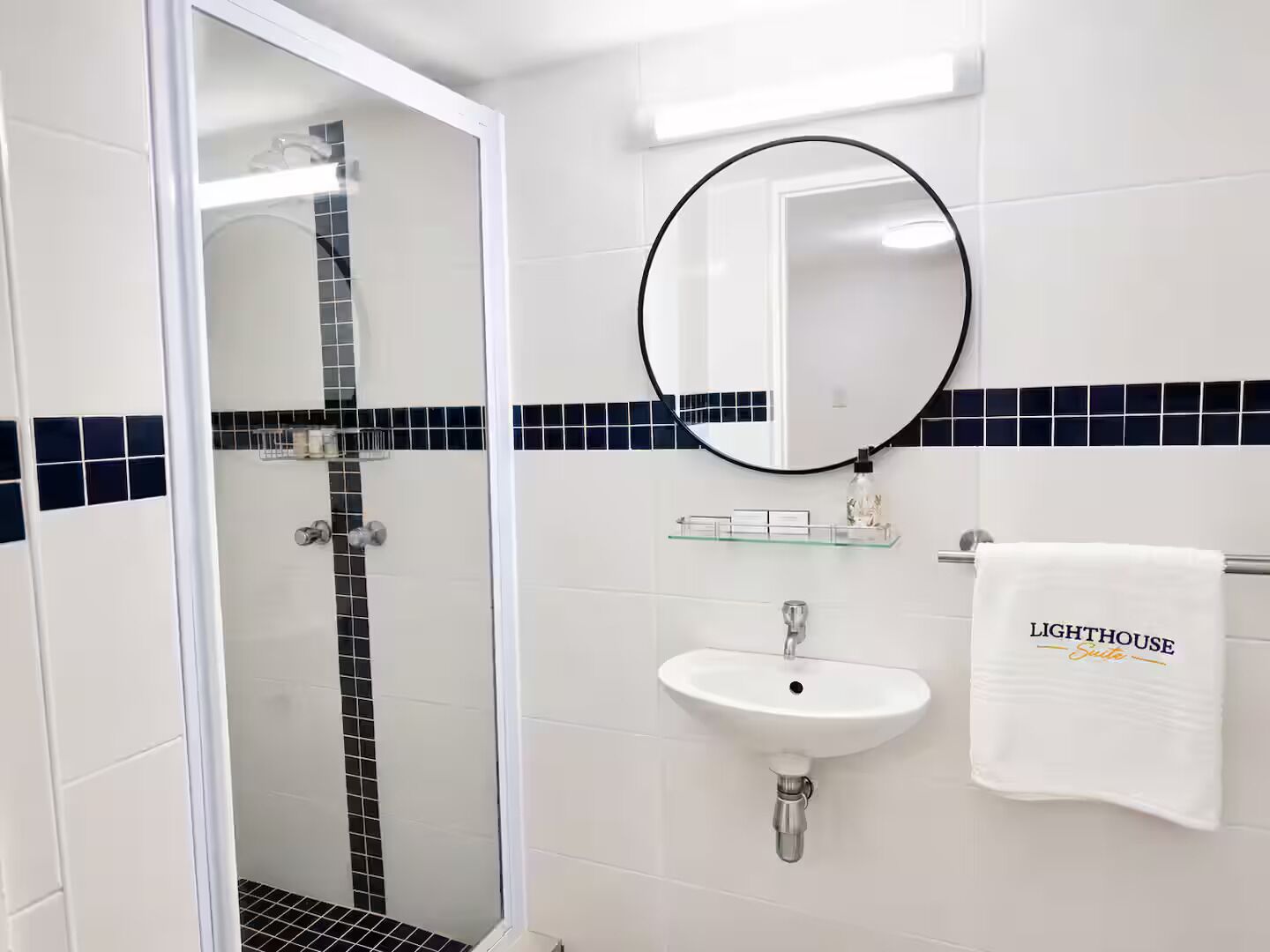 Baño con ducha de vidrio, lavamanos blanco y toallero con una toalla que dice 'LIGHTHOUSE Suite'.