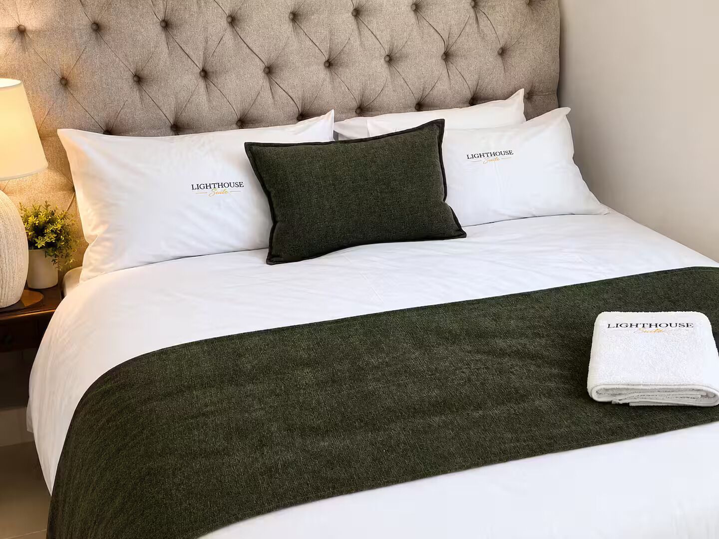 Un lit bien fait, avec une tête de lit capitonnée, du linge de lit blanc et un plaid vert foncé.