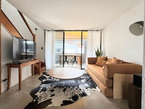 A modern living room with a sofa, a coffee table, a TV on a stand, and a dining area with chairs and a table.
