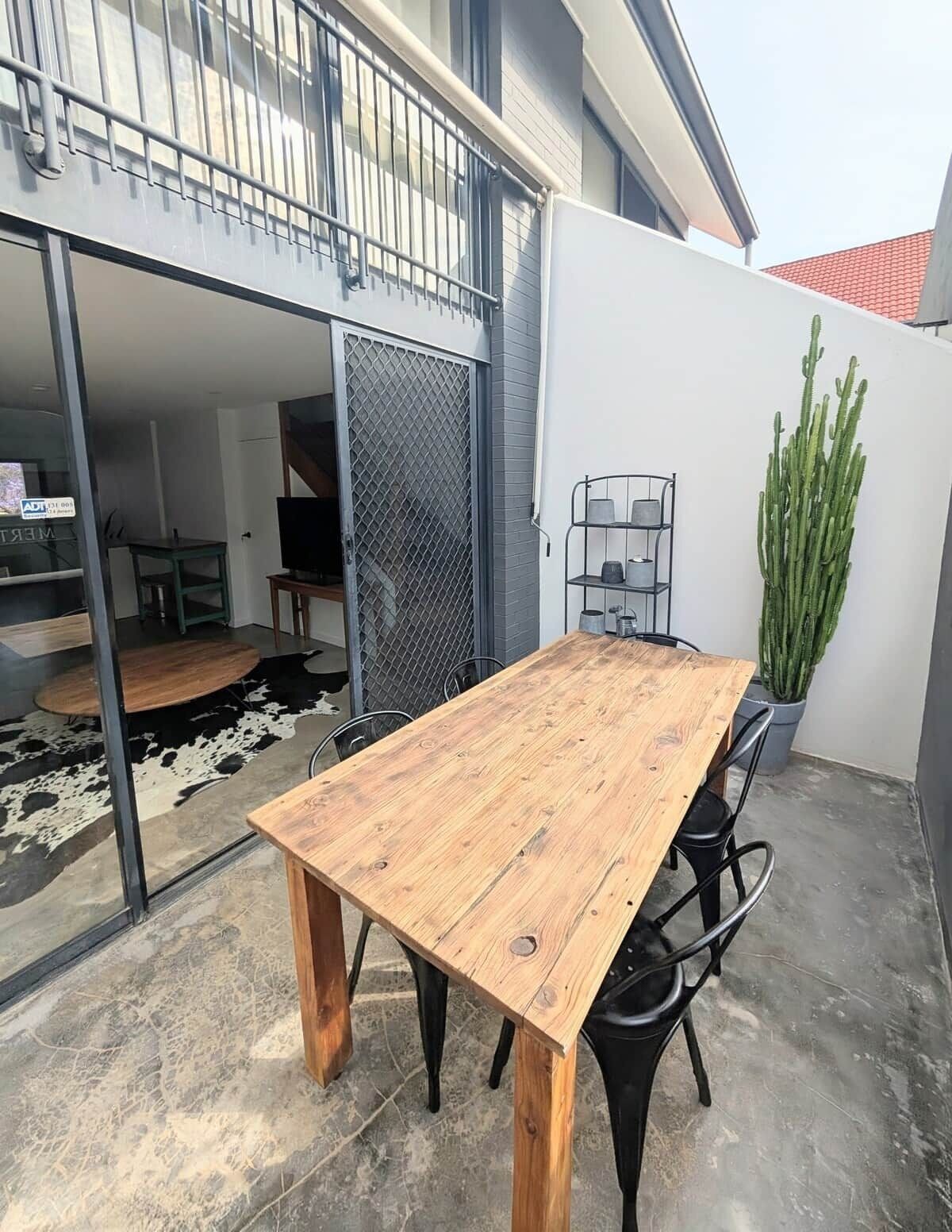 An outdoor patio with a wooden table and chairs, a metal chair, a potted cactus, and a metal shelf with containers.