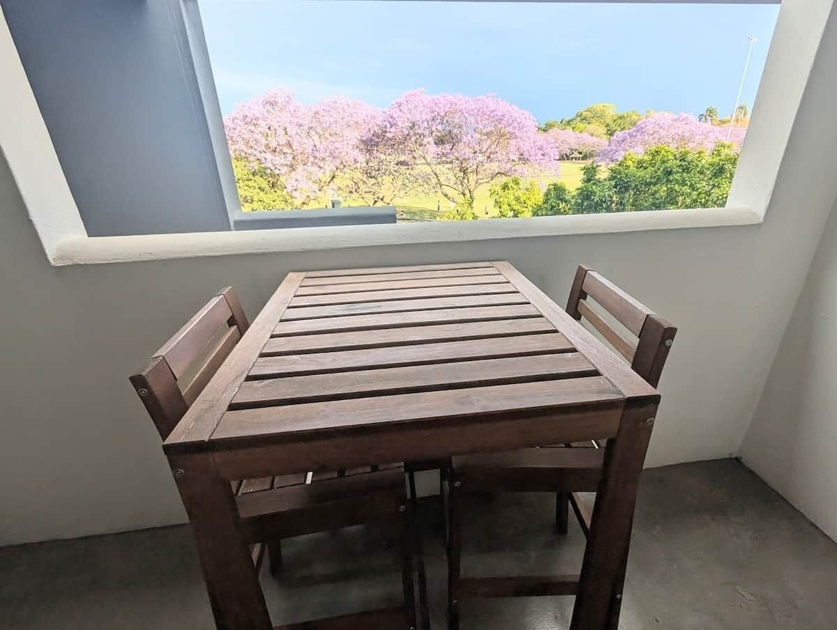 A wooden table with four chairs in a corner, positioned under a window with a view of trees.