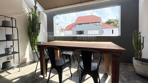 Un coin repas avec une table et des chaises, une grande fenêtre donnant sur un immeuble et des plantes en pot.