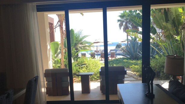 A room with a view of a pool and palm trees, a table, and a chair. - Posh 2-bedroom condo in charming Cabo with AC, kitchen and 2.5 baths.Ocean view. (Cabo San Lucas)