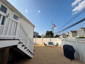 A residential backyard with a white house, a flagpole with an American flag, a covered grill, and outdoor furniture. - Perfect Peaceful Shore Getaway! Close to beach, private yard, off street parking (Mantoloking)