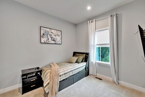 Ein Schlafzimmer mit einem Bett, einem Nachttisch, einer Kommode, einem Fenster mit Vorhängen und einem Wandbild.