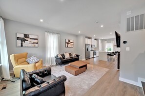 Ein modernes Wohnzimmer mit Sofa, Sessel und Couchtisch, eine Küche mit Geräte und ein Essbereich mit Stühlen.