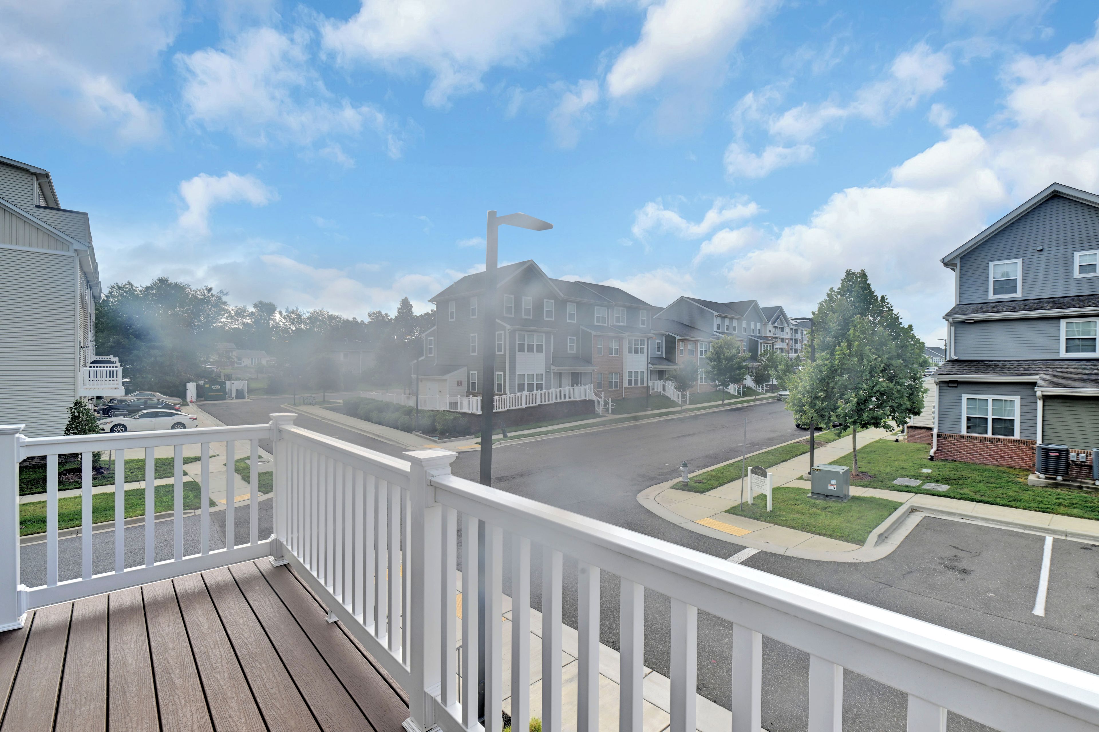 Une rue résidentielle bordée de maisons, un ciel dégagé et une terrasse en bois avec une balustrade blanche.