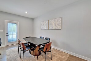 Ein Essbereich mit einem rechteckigen Tisch, der von sechs Stühlen umgeben ist, einem großen abstrakten Gemälde an der Wand und einer Tür mit Jalousien.