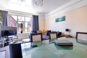 A modern living room with a glass dining table, a sofa, a TV, and a large window with curtains. - Charming Apartment in Rhos on Sea (Colwyn Bay)