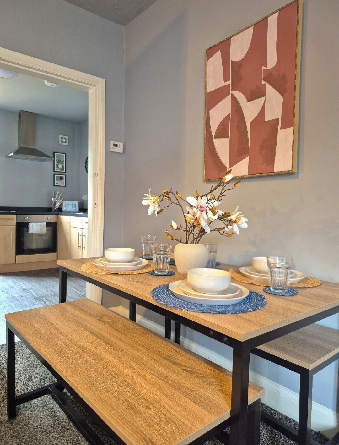 Un coin repas avec une table en bois dressée pour quatre personnes, un vase rempli de fleurs et un tableau abstrait moderne accroché au mur.